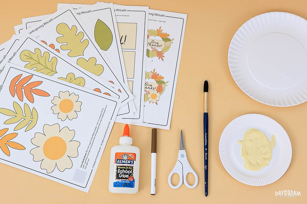 Supplies for paper plate Thanksgiving wreath craft including free printable templates, glue, scissors, marker, paintbrush, paper plates, and creamy paint.