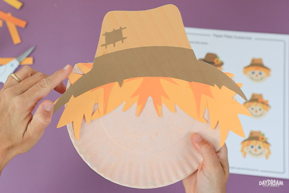 Paper plate scarecrow craft with jagged straw hair tucked under a brown harvest hat, trimming excess hair sticking out