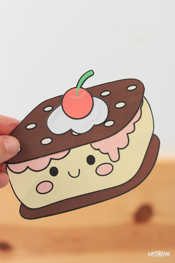 Easy ice cream craft for kids featuring a printable ice cream sandwich paper craft with chocolate cookies, pink drip icing, and cherry topping.